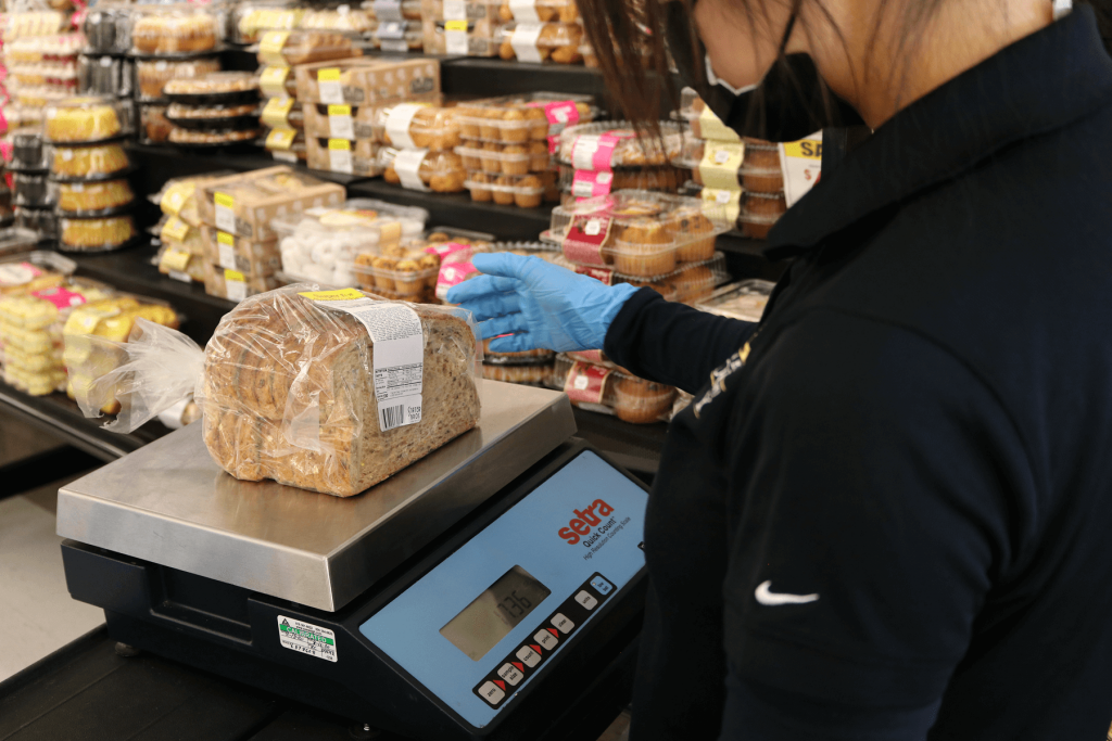 An AWM Agricultural/Standards Officer inspects the weight and price using a small scale on a loaf of bread at a supermarket.