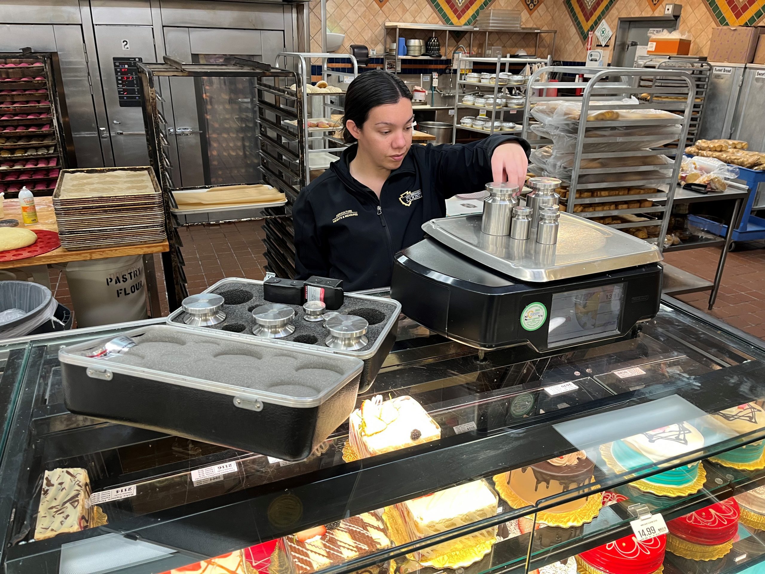 A female Agriculture/Weights & Measure Standards Officer weighs a bakery consumer scale with large steel commercial weights.