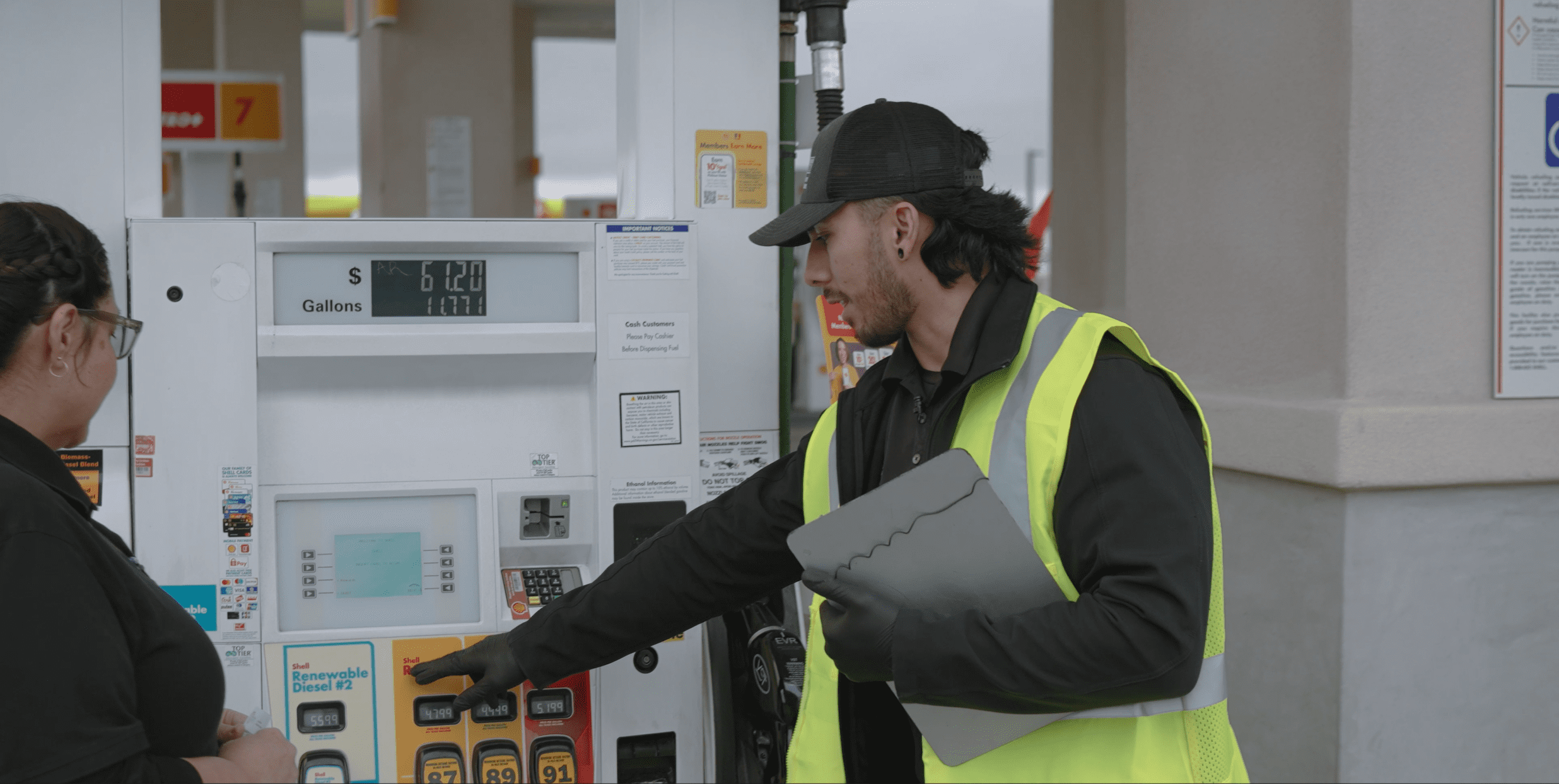 A man dressed in black uniform with ball cap and fluorescent yellow vest holds a clip board and points to a fuel pump dispenser at a gas station while a woman wearing glasses looks where he points.